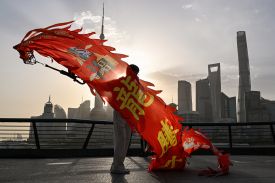 A person on a waterfront promenade holds a long, vibrant red dragon streamer with yellow characters painted on it. In the background, the sun sets behind a dense skyline of modern skyscrapers.