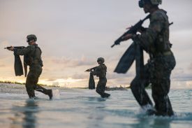 Several military personnel in camouflage gear and helmets run through shallow ocean water toward a beach at dusk. They carry rifles and have large diving fins hanging from their gear. The sky is overcast with a faint glow on the horizon.