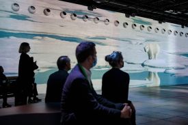 A group of people, some wearing face masks, sit in a darkened room facing a large floor-to-ceiling projection of a polar bear standing on an ice floe in an Arctic landscape.