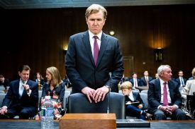 Elbridge Colby stands behind a chair with a microphone, about to sit down. He wars a dark business suit and a serious expression. Behind him are seated officials, aides, and one child.