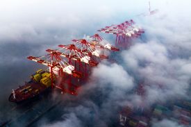An aerial view shows a row of massive red and white gantry cranes at a shipping port, partially obscured by thick, swirling fog. On the left, a large cargo ship loaded with yellow containers is docked beneath the cranes. Stacks of multicolored shipping containers are faintly visible through the mist in the lower right corner.
