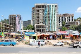 The shells of midrise residential buildings covered in scaffolding rise up against a clear blue sky. In the foreground are colorful tents and booths of vendors lining a street, where cars and vans pass by.