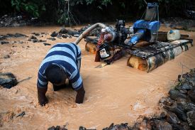 A man in a blue striped shirt reaches his hands into a muddy river; behind him is a makeshift dredger.