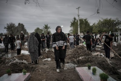 A woman in a black cloak and head covering walks through a dirt area with mounds of earth and some greenery in pots atop gravesites.