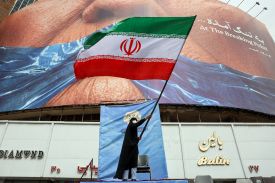 A person wearing a black chador and a face mask stands on a small blue stage, holding a large Iranian flag that waves in the wind. Behind them is a massive billboard featuring a close-up of a person’s face and text in English and Farsi. White storefronts with gold and red lettering are visible at the bottom.
