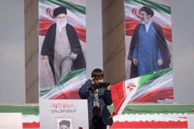 A young boy stands in front of a white sign that says "NO WAR on children," holding a small Iranian flag and taking a photo with a smartphone. In the background, two large vertical banners display portraits of Iranian leaders against a pale sky.