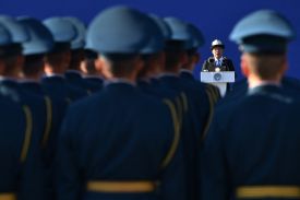 A man in a suit and a traditional white hat speaks at a white lectern. He is viewed from behind a formation of numerous uniformed personnel in blue caps, who are blurred in the foreground. The speaker is positioned in the distance against a solid blue background.