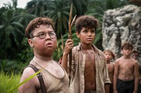 A medium close-up of two young boys standing in a tropical jungle. The boy in the foreground wears round glasses and a tan undershirt with suspenders. The boy behind him has curly hair, wears a tan button-down shirt, and holds a long, forked wooden staff. Other boys are visible in the blurred background.