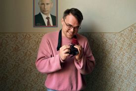 A man in a pink shirt, holding a camera, smiles in front of a poster of Putin.