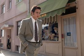 A man in a grey suit with his hands in his pockets walks down a street, with a storefront in the background.