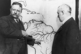 In this black-and-white photo, Teddy Roosevelt points to the middle of South America on a map as another man looks at the map from the other side of it. Both men wear suits, ties, and wire-frame glasses.
