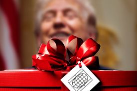 A photo illustration shows a close-up shot of a red gift box topped with a large, shiny red satin bow. Attached to the ribbon is a white square gift tag featuring a black line-art icon of a globe within a rectangular frame. In the soft-focus, blurred background, the face of a smiling person is visible behind the gift.