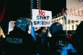 Police surround a protester holding a sign that reads "No Sport Washing."
