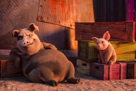 A very large, greyish-brown pig smirks and reclines comfortably against a wooden crate. To the right, a much smaller pink piglet sits upright on a stack of colorful wooden crates, looking toward the larger pig with a smile.