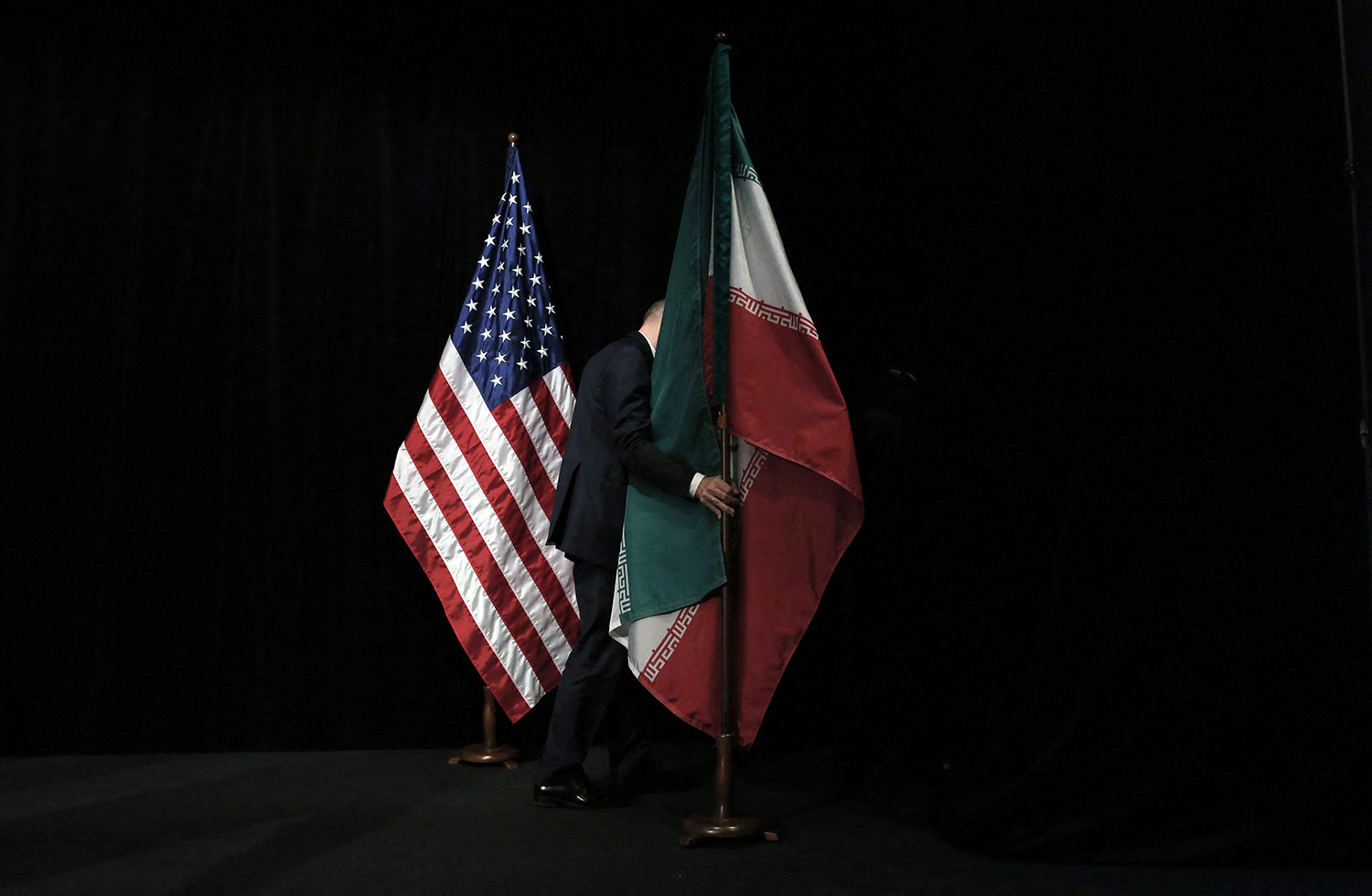 The American and Iranian flags stand side-by-side on wooden poles against a black background. A person in a dark suit is partially visible behind the Iranian flag, reaching out to adjust or hold the flagpole, their face obscured by the fall of the flag.