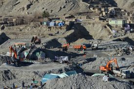 Workers and construction equipment are seen from a distance in a moonlike, cratered landscape torn up by mining activity. On a ridge in the distance are a few tents and simple, single-story buildings.