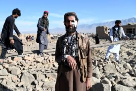 A bearded man with a bandage high up on his cheek and one arm in a makeshift sling made from a scarf stares at the camera. Behind him, three other men climb around a pile of rubble, seemingly mostly stone and concrete, from a demolished building.