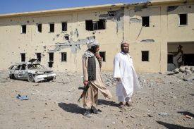 Two men stand in front of a damaged two-story building, which is covered with pockmarks. The destroyed shell of a car sits amid rubble in front of the building. One man holds an automatic rifle and has his hand on his chin as he speaks to the other man.