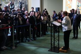 Bachelet is seen in profile as she stands at a podium, gesturing with both hands as she speaks into a microphone. Dozens of people stand clustered in front of her, some pointing cameras or microphones in her direction.
