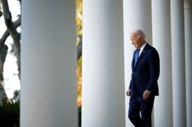 Biden walks between massive white columns outside of the White House. He wears a dark suit and a blue tie, and his head is tilted down toward the ground.