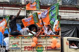 People sit in the open bed of a truck decorated with orange balloons and orange-and-green BJP party flags. A man perched on the wall of the truckbed waves his fist in the air, and an older man facing the camera waves another BJP flag.