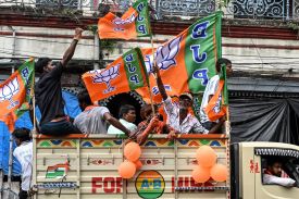 People sit in the open bed of a truck decorated with orange balloons and orange-and-green BJP party flags. A man perched on the wall of the truckbed waves his fist in the air, and an older man facing the camera waves another BJP flag.