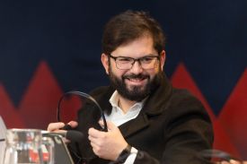 Boric smiles as he holds a pair of headphones. He is a man in his late 30s wearing a white shirt and black coat in front of a blue and red background.