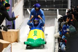 One man raises his arms in celebration as he sits inside a green and yellow bobsleigh. Another man rises up behind him.