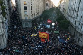 Thousands of people crowd in an intersection between two city streets, with the crowds stretching down the streets beyond view. Many hold banners or smaller signs.