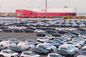A wide shot shows a large parking lot full of hundreds of neatly lined up new cars and SUVs. In the background is a harbor with a massive carrier ship looming against a hazy gray sky.