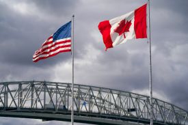 The U.S. and Canadian flags flutter in the wind. Behind them is the Blue Water Bridge, which connects Michigan in the United States and Ontario in Canada.