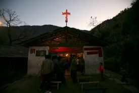 Dozens of people throng around the front door of a small church building beneath a dim sky. The people are cast in shadow, but warm light glows from within the church and from a small light-up cross on its roof.