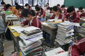 High school students in matching maroon and gray jackets sit in a classroom and study at desks piled high with stacks of books, notebooks, and papers at their sides.