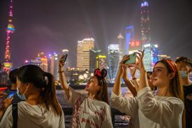 Three women lift up their phones to take flash photographs of something seen off-camera, lit by spotlights and neon from a shining cityscape beneath the night sky across the river behind the women. Other people mill about at the edge of the frame.