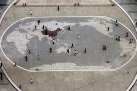 Two dozen or so scattered pedestrians walking on a large plaza are seen from overhead. There is a giant world map embedded in the pavement, with China highlighted in a dark red and all the other countries in pale gray.