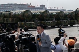 A man in a gray suit stands in the foreground of four camouflaged ballistic missiles on trucks.