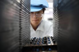 A worker wearing glasses and a blue cap looks through a piece of equipment in a workshop.