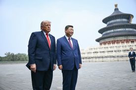 Two men in dark suits stand side-by-side in a large, paved outdoor courtyard. The man on the left wears a bright red tie and looks off to the side with a serious expression. The man on the right wears a burgundy tie and has a slight smile. In the background, a large, multi-tiered circular traditional building with a blue roof and ornate details stands under a pale sky. A person in a dark uniform is visible in the far distance to the right.