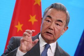 Wang, wearing a gray suit and tie, gestures with one hand bent into a claw-like shape while speaking in front of a Chinese flag and a bright blue background.