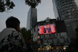 A woman is seen from behind with her neck craned to look at a large screen affixed to a four-story building with big windows. On the screen, Trump and Xi stand side by side and shake hands.