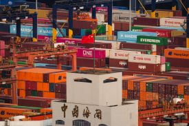 A red-and-yellow Chinese flag flies from a flagpole on the top of a simple building of about two stories tall. The building is surrounded on all sides by colorful shipping containers stacked in various configurations.