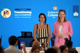 Van Veldhoven and Velez stand in front of a blue wall showing the name and logo of the conference, both smiling slightly toward out-of-focus audience members in the foreground. Veldhoven is wearing a pink pantsuit and Velez is wearing a black and white striped sleeveless pantsuit.