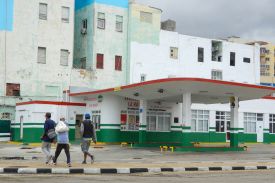 Three people walk away from the camera in front of a gas station building with blinds drawn over the windows. One man carries a sack over his back as he walks.