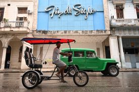 A bicycle taxi with a red roof and a green car drive along a street in Havana.