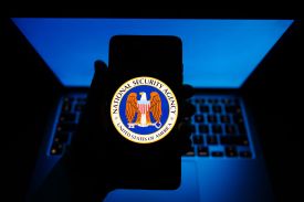 A hand in a dim room shows a smartphone screen with the U.S. NSA seal on its screen, showing an eagle with the American flag and the agency name on the circular rim of the seal. The hand is silhouetted by dim blue light from a keyboard behind it.