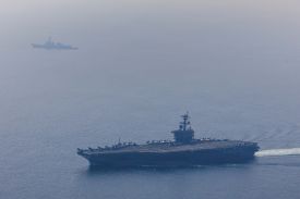 A massive aircraft carrier cuts across the water through a light fog. another ship is visible farther in the background.