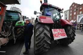 A man stands between two tractors, one green and one red, with tires as tall as his shoulders. A spray painted sign at the front of the red tractor says "No fuel, no food." The sky overhead is gray, and midrise buildings are visible across the street.
