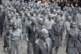 A group of dozens of people covered in gray clay walk in a mass crowd with blank expressions on their faces, arms limp at their sides. Many wear business suits and other formal clothing beneath the caked-on clay.