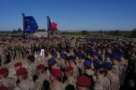 Macron is seen from a slight distance over the heads of soldiers in combat fatigues standing in neat lines beneath a clear blue sky as they listen to him. Macron himself stands at a podium in a black suit and dark tie. French and EU flags fly behind him.