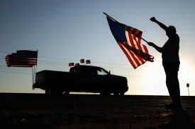 A silhouette of a person standing against a bright, hazy sky, holding a large American flag and raising a clenched fist. In the background, a silhouette of a pickup truck is parked with several more American flags attached to its bed.